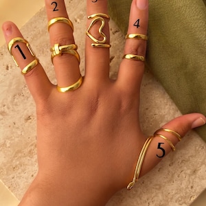 Könnte beinhalten: Eine Sammlung goldfarbener Ringe an den Fingern, nummeriert von 1 bis 5. Die Ringe weisen verschiedene Designs auf, darunter ein langer vertikaler Stab, ein Knoten, eine Herzform, einfache Bänder und ein mehrfach gewickelter Stil am Daumen.