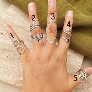 Puede incluir: Cinco anillos de metal plateado usados en los dedos, numerados del 1 al 5. El anillo 1 presenta un diseño de doble bucle, el 2 es una banda de múltiples espirales, el 3 es un óvalo alargado, el 4 es una espiral con extremos planos y el 5 es una espiral.