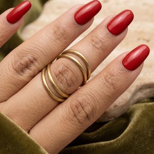 May include: A set of three thin, smooth, gold-toned metal rings worn stacked on a finger. The rings feature a simple, rounded band design with a matte finish. The metal has a warm, yellow-gold hue that contrasts with the bright red nail polish.