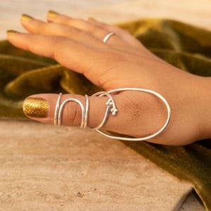 May include: A silver-toned metal finger splint or ring featuring a continuous wire design that wraps around the thumb. The structure includes a series of curved loops and a larger oval frame, accented with small spherical beads at the terminal ends.