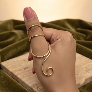 May include: A gold-toned metal ring featuring a multi-band design that wraps around the thumb. The piece consists of two simple circular bands at the top and a larger, open spiral-shaped section that extends downward along the thumb.