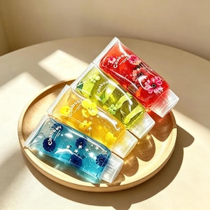 Può includere: Quattro tubetti trasparenti riempiti di gel colorato, ognuno con il marchio Chaboky e piccoli accenti floreali. I tubetti sono blu, gialli, verdi e rossi e contengono ciascuno petali di fiori distinti e forme a stella.