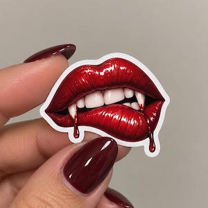 May include: A die-cut sticker featuring a pair of glossy, vibrant red lips biting the lower lip, revealing sharp white vampire fangs. The design includes realistic red liquid dripping from the corners of the mouth, set against a clean white border.