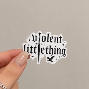 May include: A die-cut sticker featuring the black text violent littlething in a gothic font. The letter i in violent is replaced by a vertical sword illustration. A small black dragon silhouette and several four-pointed stars surround the text.
