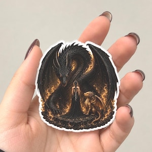 May include: A die-cut sticker featuring a large black dragon with glowing orange eyes and wings, standing behind a figure in a long dark dress, with a smaller golden dragon beside them, all surrounded by swirling orange flames on a dark background.