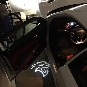 Puede incluir: Un coche blanco con la puerta del conductor abierta, proyectando en el suelo un logotipo blanco brillante con forma de cabeza de felino. El interior muestra un salpicadero negro, un volante y detalles rojos en los paneles de las puertas.