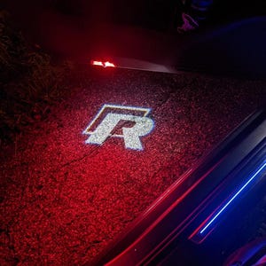 Könnte beinhalten: Ein hellweißes, projiziertes Lichtlogo des Buchstabens R mit blauem Umriss, das auf eine dunkle Asphaltfläche geworfen wird. Die Projektion wird von einer Fahrzeugtür beleuchtet und bildet einen scharfen Kontrast zum roten Umgebungslicht.