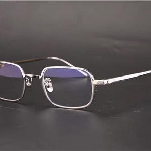 May include: A pair of rectangular eyeglasses featuring a thin, polished silver-tone metal frame. The lenses are clear with a subtle blue-tinted reflection. The temples are slender and metallic, connecting to the frame front with small hinges.