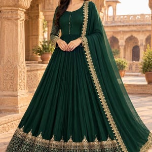 May include: A dark green floor-length gown with long sleeves, featuring a round neckline with a vertical row of small buttons. The hem and cuffs are adorned with intricate gold embroidery and a scalloped edge. A matching sheer dupatta is draped over one shoulder.