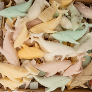 May include: A collection of small, whale-shaped soap bars in pastel shades of cream, pale yellow, light pink, and mint green, arranged inside a cardboard box lined with floral-patterned tissue paper and filled with shredded paper filler.