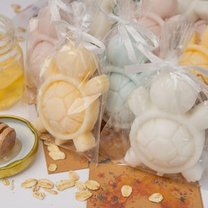 May include: Several turtle-shaped soaps in clear plastic bags tied with white ribbons, accompanied by a small glass jar of golden honey, a wooden honey dipper, and scattered oat flakes. The soaps are pastel yellow, white, and light blue.