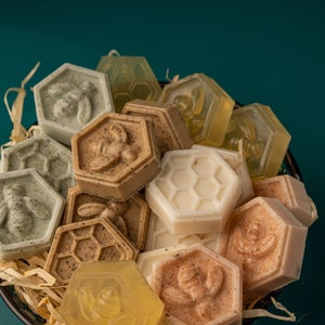 Op de afbeelding: Een verzameling handgemaakte zeepjes in verschillende vormen en kleuren, waaronder bruin, beige en wit. De zeepjes zijn gerangschikt in een mand met stro en hebben bijen- en honingraatontwerpen.