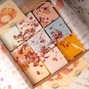 Op de afbeelding: Een doos handgemaakte zepen in verschillende kleuren, waaronder roze, blauw en geel, elk bedekt met gedroogde bloemblaadjes. De zepen zijn gerangschikt in een kartonnen doos, met een decoratieve kaart met een cartoon illustratie.