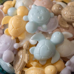 May include: A collection of small, moulded turtle-shaped soaps in various pastel colours including light blue, yellow, lavender, and tan. Some turtles feature a heart-shaped shell pattern, while others display a star-patterned shell or a speckled texture.