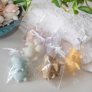 May include: Four individual clear plastic bags each containing a turtle-shaped soap. The soaps are light blue, white, tan with speckles, and yellow. Each bag is tied at the top with a thin white ribbon. A bowl of additional soaps is visible.