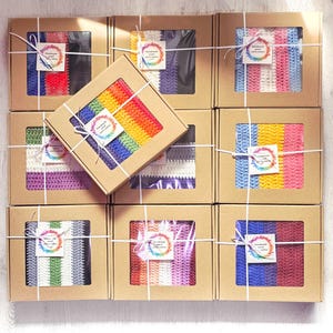 May include: Nine square, brown cardboard boxes with clear windows, each containing a colorful, crocheted item. The boxes are tied with white string and have a small, round tag. The crocheted items feature stripes of various colors, including rainbow, blue, and purple.