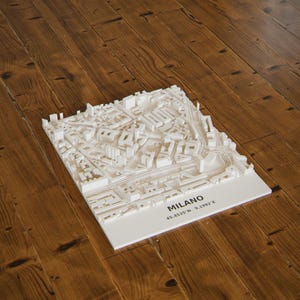 Può includere: Un modello architettonico 3D bianco della città di Milano, caratterizzato da blocchi di edifici dettagliati e layout stradali. La base include il testo stampato MILANO e le coordinate 45.4535°N 9.1992°E in inchiostro nero sulla superficie.
