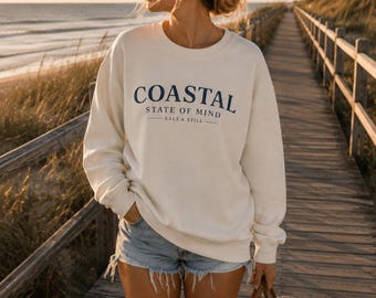Könnte beinhalten: Ein cremefarbenes Sweatshirt mit Rundhalsausschnitt und dem dunkelblauen Schriftzug COASTAL STATE OF MIND SALT & STILL auf der Brust, kombiniert mit hellblauen Jeansshorts und einer geflochtenen Strohtasche mit dunkelblauen Querstreifen.