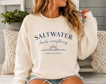 Könnte beinhalten: Ein cremefarbenes Sweatshirt mit Rundhalsausschnitt und dunkelblauem Text, der SALTWATER heals everything lautet, ergänzt durch eine Grafik einer aufgehenden Sonne über Wellen und den Text SALT & STILL, der unter dem Design auf der Vorderseite steht.