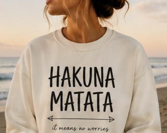 Könnte beinhalten: Ein cremefarbenes Sweatshirt mit Rundhalsausschnitt und dem schwarzen Schriftzug HAKUNA MATATA in einer fetten, abgerundeten Schriftart. Unter dem Text befindet sich ein horizontaler Pfeil, gefolgt von dem kursiven schwarzen Text it means no worries.