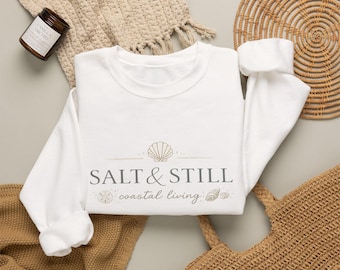 Könnte beinhalten: Ein weißes Langarm-Sweatshirt mit einem Grafikdruck in Beige und Grau. Das Design zeigt ein Muschelsymbol über dem Text SALT & STILL und dem in Schreibschrift gehaltenen Text coastal living, flankiert von kleinen Abbildungen einer Sanddollar-Muschel.