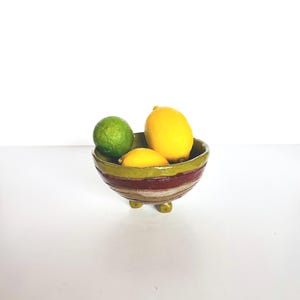 May include: A small, round, three-legged ceramic bowl with a glossy finish, featuring horizontal bands of olive green, deep maroon, and cream. The bowl contains one bright green lime and two vibrant yellow lemons resting inside the basin.