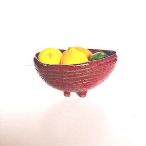 May include: A deep, textured magenta ceramic bowl with horizontal ridges and three small feet. The bowl is filled with two bright yellow lemons and one dark green lime, showcasing a contrast between the vibrant fruit and the dark red vessel.