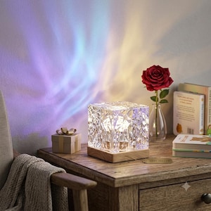May include: A square, faceted crystal lamp sits on a light wood base, casting colorful blue and yellow light patterns on the wall. The lamp features a textured, geometric surface that refracts light, creating a dynamic visual effect in the room.