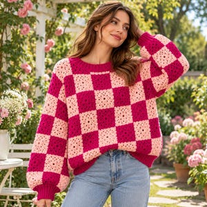 May include: A long-sleeve sweater featuring a checkered pattern of alternating bright magenta and light pink crochet granny squares, with a ribbed crew neckline, cuffs, and hem, paired with light blue denim jeans.