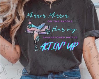 May include: A charcoal gray t-shirt featuring a graphic of a colorful western saddle with turquoise and pink accents. The text reads: Mirror Mirror ON THE SADDLE Hear my RHINESTONES RATTLE ACT IN' UP. The design uses bright cyan and purple lettering.