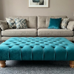May include: A large, rectangular, teal velvet ottoman featuring deep button tufting across the top surface. The piece is supported by four short, rounded, light-coloured wooden feet, providing a low-profile base for this furniture item.