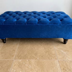 May include: A rectangular ottoman upholstered in vibrant blue velvet fabric featuring a deep button-tufted top surface. The piece is supported by four short, dark brown, turned wooden legs that provide a stable base for the furniture item.