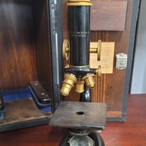 May include: A vintage Bausch & Lomb Optical Co. microscope with a black metal body and brass accents, featuring a rotating nosepiece with three objectives, a square stage, a circular mirror at the base, and a wooden storage case in the background.