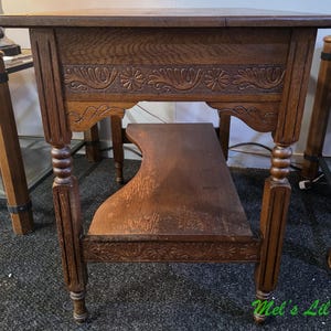May include: A brown wooden side table featuring a rectangular top, a lower shelf with a curved edge, and four legs with turned detailing. The apron of the table is decorated with carved floral and leaf patterns. The text Mel's Lil Bits is visible.