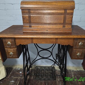 Può includere: Un tavolo da cucito vintage in rovere con coperchio in legno, due piccoli cassetti su ciascun lato con maniglie ad anello in ottone e una base a pedale in ghisa nera. Il tavolo presenta un piano rettangolare e un grembiule smerlato.