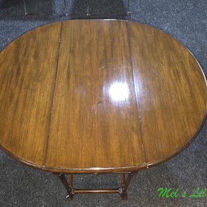 May include: A round, brown wooden drop-leaf table with a polished, smooth surface showing natural wood grain patterns. The table features a central fixed section flanked by two hinged leaves that can be folded down, supported by a wooden frame.