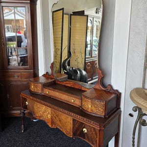 May include: A wooden vanity table featuring a large, arched mirror with a decorative top. The piece includes a central drawer, two side drawers, and two lower drawers, all accented with light-colored round knobs and intricate wood grain patterns.
