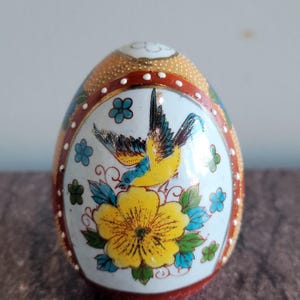 May include: A decorative egg featuring a hand-painted scene of a yellow and blue bird perched above a large yellow flower. The egg has a white background, a red border with white dots, and a textured gold top. The text Mets Lil Bits is visible.