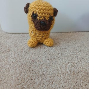 May include: A small, handmade crochet pug figurine crafted from golden-yellow yarn. The figure features a dark brown muzzle, matching dark brown ears, and two small, shiny black plastic bead eyes. It has a rounded body and two small feet.