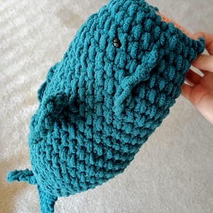 May include: A teal-colored whale plush toy crafted from chunky, textured yarn. The whale features a rounded body, small side fins, a tail, and a single visible black bead eye. A small portion of a light peach-colored fabric is visible at the mouth.