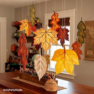 May include: A hanging mobile featuring multiple cutout leaves in shades of yellow, orange, red, brown, and green. The leaves are suspended by thin white strings above a wooden rectangular box filled with colored pencils and a small woven basket.