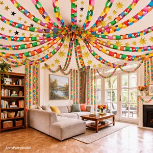 May include: A living room decorated with colorful paper chain garlands hanging from the ceiling in a radial pattern, interspersed with gold and black star cutouts. The room features a beige sectional sofa, a wooden coffee table, and a bookshelf.