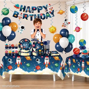 May include: A space-themed party decoration set featuring a Happy Birthday banner, dark blue tablecloths with rocket and planet prints, balloons in navy, white, and orange, hanging spiral decorations, and paper plates with the text 3.2.1 BLAST OFF!
