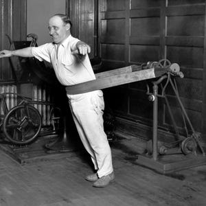 May include: A vintage mechanical exercise machine featuring a wide, horizontal belt wrapped around the waist of a person wearing a light-coloured short-sleeved shirt and trousers. The device includes a metal frame, pulleys, and a motor mechanism.