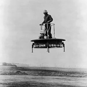 May include: A black and white photograph of a circular, disc-shaped flying platform hovering above the ground. The vehicle features a central standing area with a metal railing, landing struts, and the text USN-ONR printed on the side.