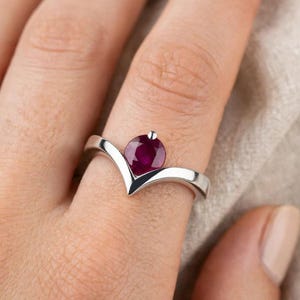 Könnte beinhalten: Ein silberfarbener V-förmiger Ring mit einem einzelnen, rund geschliffenen, tiefroten Rubin in der Mitte. Die Ringschiene hat eine glatte, polierte Oberfläche und folgt einer Chevron-Silhouette, die sich zum Stein hin verjüngt.