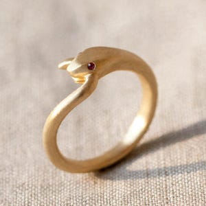 Könnte beinhalten: Ein goldfarbener Ring in stilisierter Schlangenform mit matter Oberfläche. Der Schlangenkopf ist mit einem kleinen, runden, dunkelroten Edelstein als Auge verziert, der einen dezenten Kontrast zur texturierten goldenen Oberfläche des Rings bildet.
