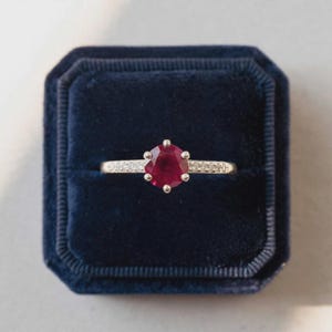 Könnte beinhalten: Ein silberner Ring mit einem runden, tiefroten Edelstein in einer sechs-Krappen-Fassung. Die Ringschiene ist mit kleinen, klaren, funkelnden Steinen besetzt, die entlang der Seiten eingelassen sind, und liegt in einer quadratischen, dunkelblauen Samtschatulle.