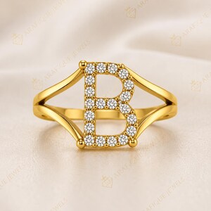 May include: A gold ring featuring a capital letter B as the centrepiece, which is encrusted with numerous small, clear, round-cut stones. The band of the ring splits into two smooth, polished gold arms that connect to the top and bottom of the letter.