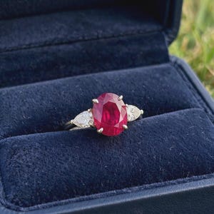 Könnte beinhalten: Ein Ring mit einem großen, oval geschliffenen, tiefroten Rubin in der Mitte, flankiert von zwei kleineren, birnenförmigen, klaren weißen Diamanten. Der Ring ist in eine polierte Metallschiene gefasst und liegt in einer dunkelblauen Schmuckschatulle.